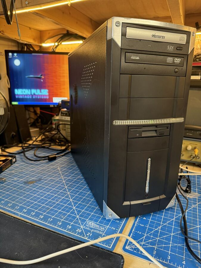 Black tower on the bench beside a monitor showing the NEON PULSE Vintage Systems wallpaper